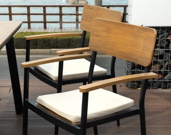 Puede incluir: Dos sillas de metal negro con respaldos de madera y cojines blancos. Las sillas tienen un diseño moderno y son perfectas para comer al aire libre.