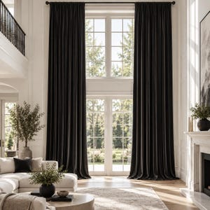 May include: Floor-to-ceiling dark charcoal curtains hanging from a black rod, covering tall windows in a room with white walls. The heavy fabric drapes in vertical folds, extending from the high ceiling down to the hardwood floor surface.