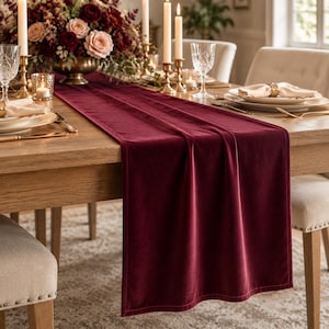May include: A deep burgundy velvet table runner with a smooth, soft texture and finished edges, draped lengthwise across a wooden dining table. The runner features a rich, solid color that extends from the center to the edge of the table.