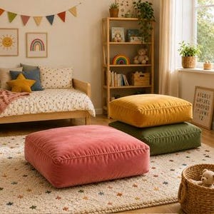 May include: Three square floor cushions in velvet fabric, featuring a pink cushion in the foreground and a stacked pair of yellow and olive green cushions behind it. These plush, low-profile seating pieces have piped edges and a soft texture.