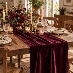May include: A deep burgundy velvet table runner draped over a wooden dining table, accompanied by a floral centerpiece with dark red flowers, tall taper candles in brass holders, crystal glassware, and gold-toned cutlery settings.