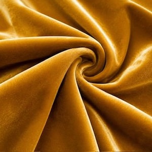 May include: A mustard yellow velvet fabric with a dense, soft pile and a smooth, reflective surface. The material is arranged in deep, swirling folds and cascading drapes that create a play of light and shadow across the rich, golden-toned textile.