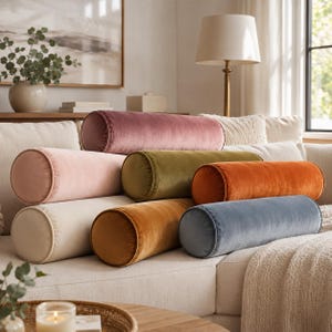 May include: A collection of seven cylindrical bolster pillows made of soft velvet fabric. The pillows are arranged in a stack and feature a variety of solid colors including dusty rose, light pink, olive green, burnt orange, cream, gold, and blue.