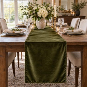 May include: A long, rectangular olive green velvet table runner draped over a wooden dining table. The runner features a soft, plush texture and finished edges, centered beneath a white floral centerpiece, crystal glassware, and candles.