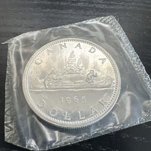 Könnte beinhalten: Eine kanadische Silbermünze im Wert von einem Dollar aus dem Jahr 1965 mit einem Kanu mit zwei paddelnden Figuren, einem Baum und dem Text "CANADA" und "DOLLAR".