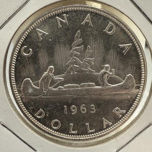 Könnte beinhalten: Eine silberne kanadische 1-Dollar-Münze von 1963. Die Münze zeigt ein Kanu mit zwei paddelnden Figuren vor dem Hintergrund von Bäumen. Das Wort "CANADA" ist oben in einem Bogen angeordnet, mit "DOLLAR" unten. Das Jahr "1963" ist zentriert.