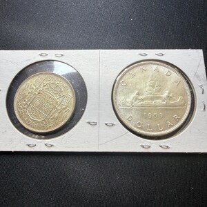 May include: Two Canadian coins, a 1953 silver dollar and a 1953 fifty-cent piece, displayed in a protective holder. The dollar features a boat scene, while the fifty-cent piece shows a coat of arms. Both coins are silver in color.