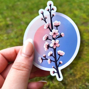 May include: A round sticker with a pink and blue ombre background featuring a white branch with pink blossoms. The sticker is perfect for adding a touch of nature to laptops, water bottles, or journals.