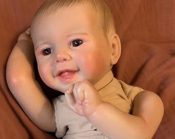 Puede incluir: Una muñeca bebé realista con una expresión sonriente, vestida con un body beige y un pañal con un diseño de Winnie the Pooh. La muñeca está acostada sobre una superficie rosa.