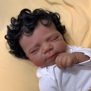 Puede incluir: Una muñeca realista con piel oscura y cabello negro rizado. La muñeca lleva un body blanco y está acostada sobre una superficie amarilla. Los ojos de la muñeca están cerrados y su mano está cerca de su rostro.