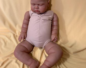 May include: A realistic baby doll with a smooth, light-toned vinyl complexion, closed eyes, and painted hair. The doll is wearing a sleeveless, light beige one-piece romper with a gathered neckline and visible creases on the arms and legs.