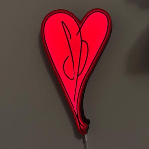 May include: A red heart-shaped neon light with a black script design. The light is plugged in and mounted on a gray wall. A musical note and a coil of cable are visible in the background.