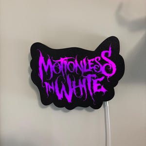 May include: A black wall light with a white cord. The light is shaped like a cloud and has the words "Motionless In White" written in a purple, gothic font.