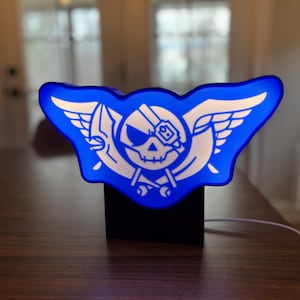 Puede incluir: Una luz nocturna azul y blanca con forma de calavera con alas. La calavera lleva un parche en el ojo y tiene huesos cruzados detrás.