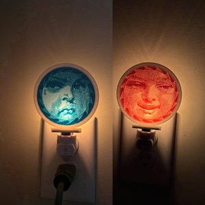 Puede incluir: Dos luces de noche enchufadas a una toma de corriente. Una luz de noche tiene un diseño de cara azul, la otra un diseño de cara roja. Las luces de noche son redondas con un borde blanco y están encendidas.