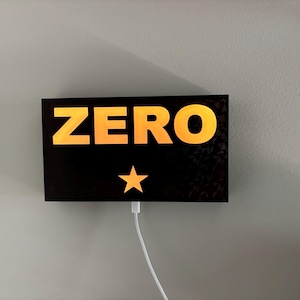 Puede incluir: Un letrero rectangular negro con la palabra "ZERO" en letras grandes de color naranja y una estrella dorada debajo. Un cable blanco sale de la parte inferior del letrero, montado en una pared gris.
