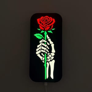 May include: A black, rectangular LED light featuring a red rose held by a white skeleton hand. The rose stem is green, and the light glows. The design is a unique decorative piece.