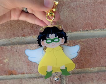 May include: A clear acrylic charm featuring a cartoon angel with black hair, green glasses, and a yellow outfit. The angel has white wings and a halo. The charm is attached to a gold-colored star-shaped clasp with a small silver bell.