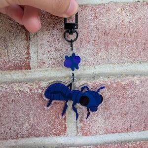 May include: A clear acrylic keychain featuring a dark blue ant illustration and a small matching blue flower charm. The keychain is attached to a black metal lobster clasp, showcasing a detailed insect design with dark eyes and segmented legs.