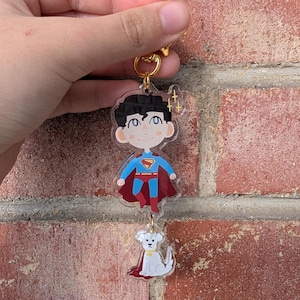 May include: A clear acrylic keyring featuring a cartoon Superman character with a red cape and blue suit, and a white dog with a red cape. The keyring has a gold-coloured ring.