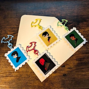 Puede incluir: Cuatro llaveros de acrílico con forma de sello postal, cada uno con una ilustración de perfil de un personaje y el texto Gotham Forever. Los llaveros tienen cierres en forma de murciélago en colores azul claro, amarillo, rojo y verde claro.