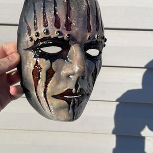 May include: A handcrafted mask with a distressed, aged appearance. The mask features a gray base with dark streaks and red accents, resembling blood. Black details outline the eyes and mouth, with small black studs. The mask is held by a hand.