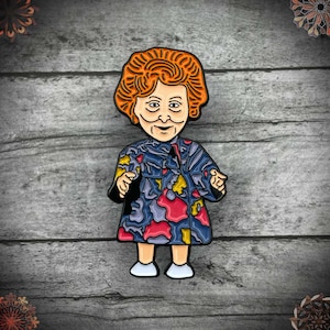 May include: Enamel pin depicting a cartoon character with orange hair and a patterned blue, red, and yellow dress. The pin has a black outline and white shoes. The background is a grey wooden surface.
