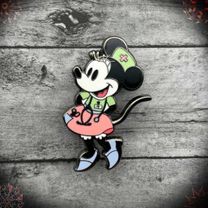 May include: A metal enamel pin featuring a cartoon mouse character dressed as a nurse. The character wears a light green top, a pink skirt with a stethoscope graphic, and a light green nurse cap with a pink cross. The mouse has black ears.