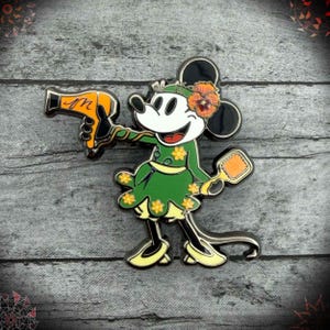 May include: Enamel pin featuring a cartoon mouse in a green dress with yellow flower accents, holding a yellow hairdryer and a small mirror. The mouse has a flower in its ear and is smiling. The pin is set against a gray wooden background.