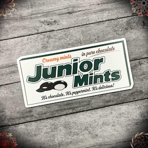 May include: A white rectangular sign with green and black lettering that reads "Creamy mints in pure chocolate Junior Mints It's chocolate. It's peppermint. It's delicious!" The sign features a graphic of three Junior Mints candies.