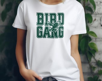 以下が含まれることがあります： 緑色のグラフィックが施された白いTシャツ。テキストには「BIRD GANG」とあり、テキストの中央には飛んでいるワシが描かれています。
