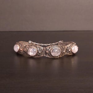 May include: A silver-toned bracelet with five oval, translucent stones. The bracelet features intricate filigree metalwork and is connected by small chain links. The stones have a soft, milky appearance, and the bracelet is displayed on a dark surface.