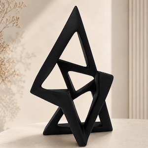May include: A matte black sculpture featuring an abstract geometric design composed of interconnected triangular and angular forms. The structure has a hollow, open-frame construction with sharp, pointed edges and a solid, dark finish throughout.