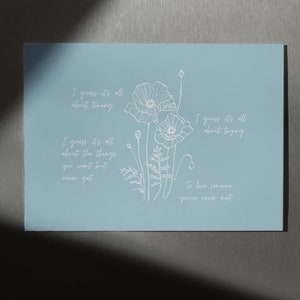 Peut inclure: Un fond bleu clair avec du texte blanc et un dessin au trait blanc de trois fleurs de pavot. Le texte dit "Je suppose que tout est une question de timing", "Je suppose que tout est une question d'essayer", "Je suppose que tout est une question des choses que tu veux mais que tu n'obtiens jamais", et "Aimer quelqu'un que tu n'as jamais rencontré".