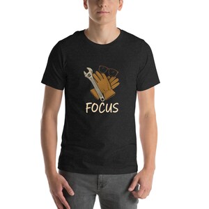 May include: A dark heather grey t-shirt with a wrench, work gloves, and sunglasses graphic. The text "FOCUS" is printed below the graphic.