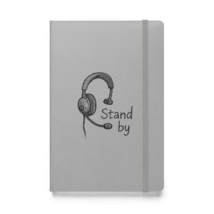 Op de afbeelding: Een grijs notitieboek met een zwart-witte illustratie van een headset en de tekst "Stand by".