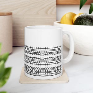 May include: White ceramic mug with a black geometric pattern of lines, zigzags, and waves. The mug has a handle and is sitting on a beige coaster.
