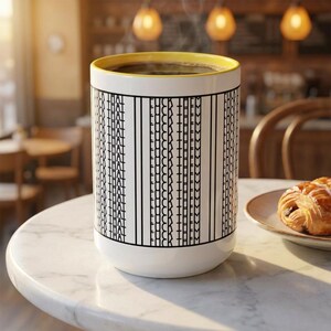 May include: White ceramic coffee mug with a yellow rim, filled with coffee. The mug features the text "FUCK THEM" and "THAT" in a repeating pattern. A croissant sits on a plate next to the mug.