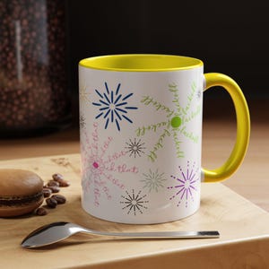 May include: White ceramic mug with a yellow interior and handle. The mug has starburst designs and the phrases "fuck that" and "fuck off". A spoon and a macaron are in the foreground. The mug is on a wooden surface.