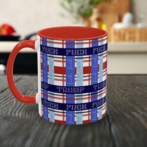 May include: A ceramic mug with a red interior and handle. The exterior features a plaid pattern in red, white, and blue, with the words "FUCK TRUMP" repeated throughout the design. The mug is sitting on a wooden surface.
