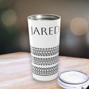 Puede incluir: Vaso aislado blanco con el nombre "JARED" en letras negras. El vaso presenta un patrón geométrico negro alrededor de la base y un borde plateado. La tapa está a un lado.