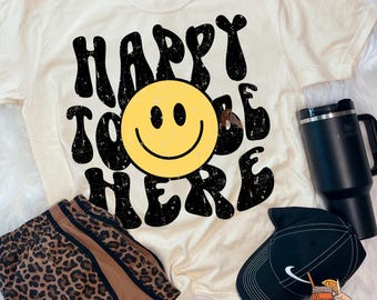 May include: Cream-colored t-shirt with the text "HAPPY TO BE HERE" and a yellow smiley face. The image also includes leopard print shorts, a black tumbler, and a black baseball cap.