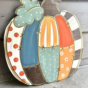 May include: A decorative wooden pumpkin wall hanging featuring a patchwork design with various patterns including orange polka dots, blue and brown stripes, white floral prints, and solid color sections, accented with black stitched details and a beaded hanger.