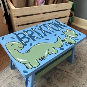May include: A light blue wooden step stool with the name "BRIXTON" painted in blue. It features a green dinosaur illustration and dinosaur footprints. The stool has a green and blue painted base and legs.