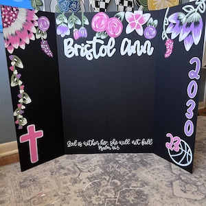 May include: A black tri-fold display board with floral and cross accents. The board features the name "Bristol Ann" in white script, the numbers "2030", and the text "God is within her, she will not fall Psalm 46:5". A basketball graphic is also present.