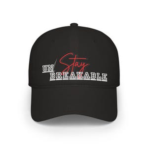 May include: Black baseball cap with the words "UN Stay BREAKABLE" in white and red lettering. The cap has a curved brim and a structured crown. The text is centered on the front of the cap.