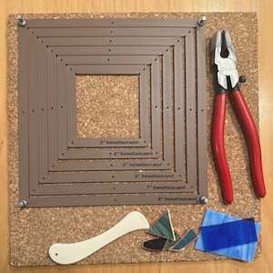 May include: A cork board with a brown stained glass layout tool, a pair of red pliers, a white plastic tool, and pieces of stained glass in various colours.