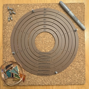 May include: A brown circular template with concentric circles marked with measurements in inches, from 3 inches to 9 inches. The template is on a cork board with a black marker and tacks.