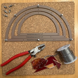 May include: A set of three brown stained glass layout tools with different sizes: 3" x 6", 4" x 5", and 3" x 10". The tools are on a cork board with a pair of red pliers, a spool of silver solder wire, and pieces of stained glass.