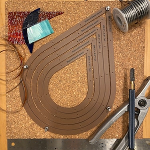 Peut inclure: Un ensemble de six gabarits en cuir brun pour des projets de vitrail. Les gabarits sont en forme de larme et sont étiquetés avec différentes tailles et hauteurs. Les gabarits sont disposés sur un panneau en liège avec une règle, un coupe-verre et du cuivre.